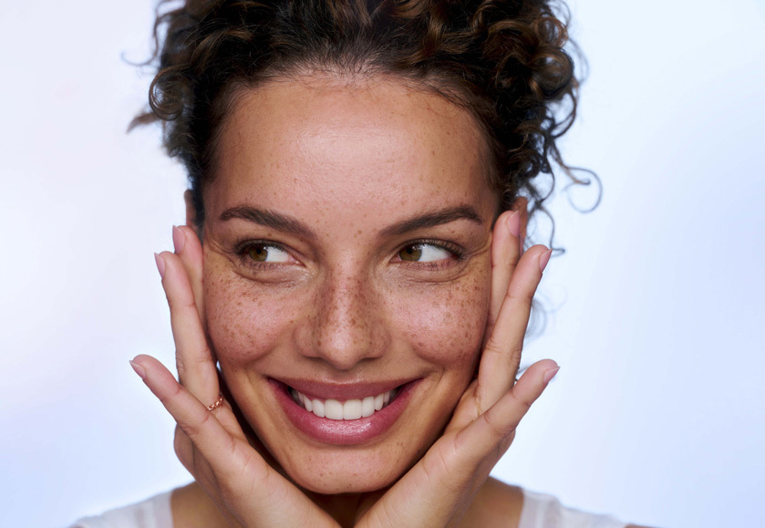 Woman with her hands holding her cheeks, smiling and looking towards her right.